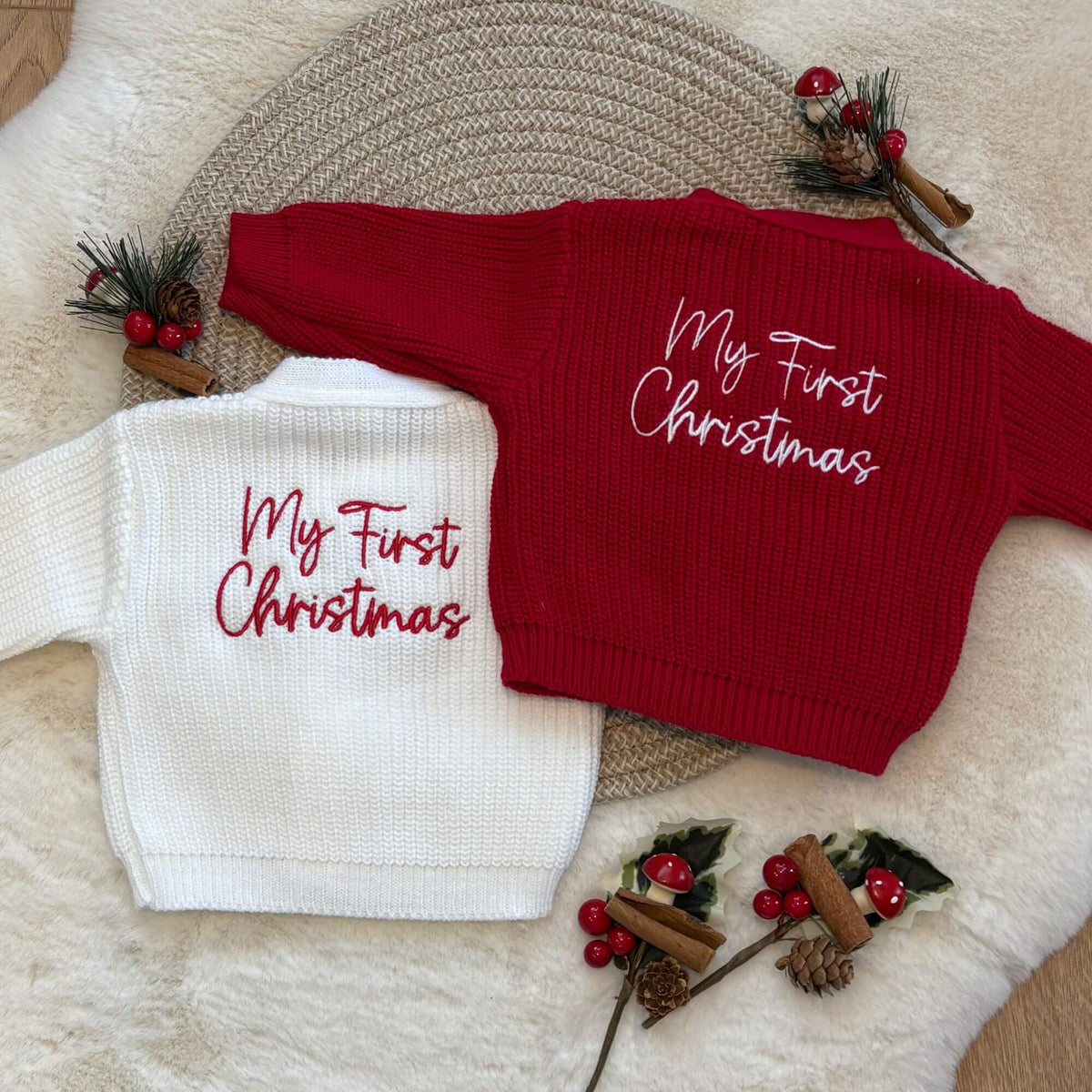 Two baby knit sweaters laid flat on a cream faux-fur surface with decorative holiday props. One sweater is white with red script text reading 'My First Christmas,' and the other is red with white script text reading 'My First Christmas.