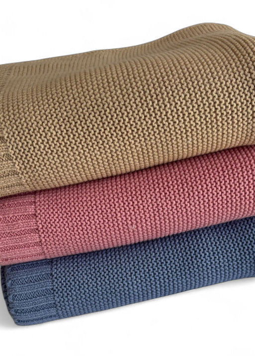 Three stacked knit blankets in beige, pink, and blue on a white background