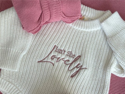 White sweater with 'Isn't She Lovely' embroidery in pink thread, with a pink knitted item in the background.