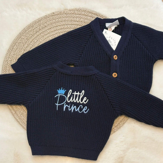 A navy blue ribbed knit baby cardigan sweater with 'Little Prince' embroidered on the back styled on a woven placemat over a soft cream background.