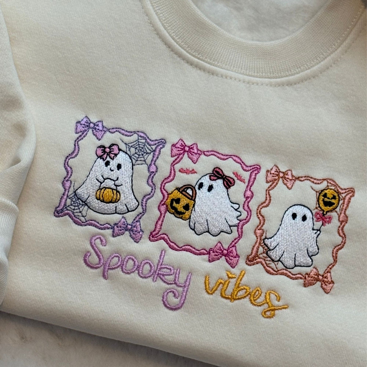 Close-up of an oat-colored sweatshirt featuring embroidered cartoon ghosts in purple, pink, and orange frames, with the text 'Spooky vibes' stitched below.