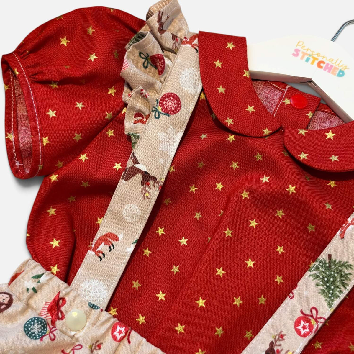 A close-up of a baby outfit set featuring a red short-sleeve top with gold star prints, beige ruffled trim, and matching beige patterned suspenders with holiday motifs, shown on a white hanger against a white background.