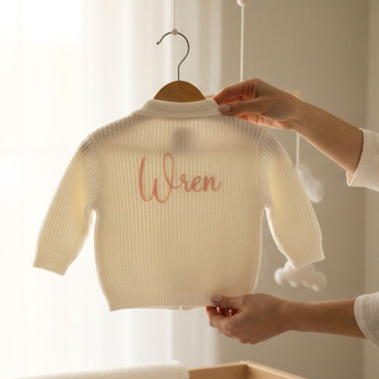 a white knit baby cardigan with babies first name embroidered on it in light pink thread