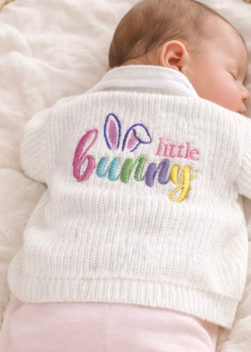 Baby wearing a white cardigan with 'little bunny' text and colorful design on a soft surface