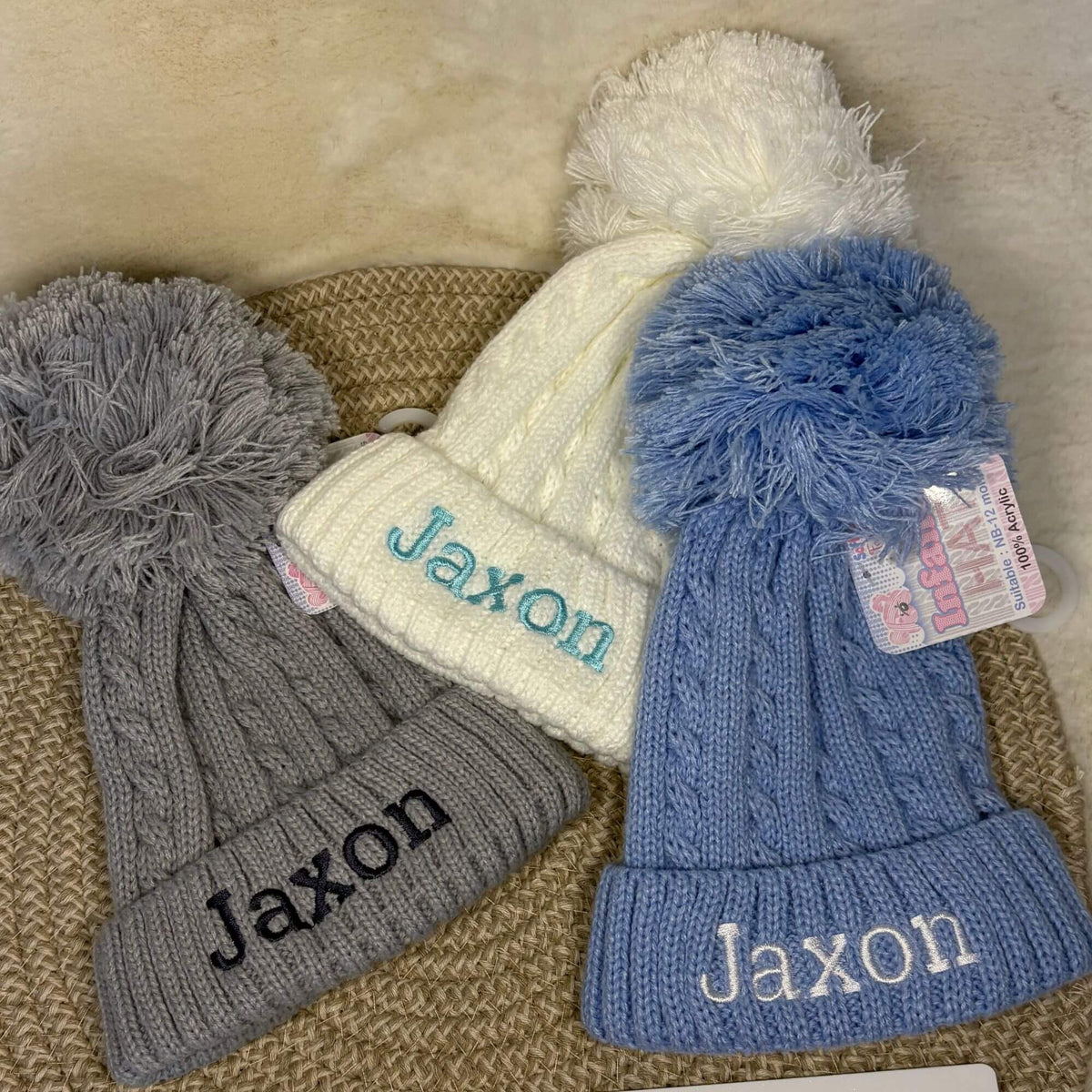 Three personalised cable-knit winter hats with pom-poms, displayed on a woven mat. The hats are gray with dark grey embroidery, white with light blue embroidery, and light blue with white embroidery.