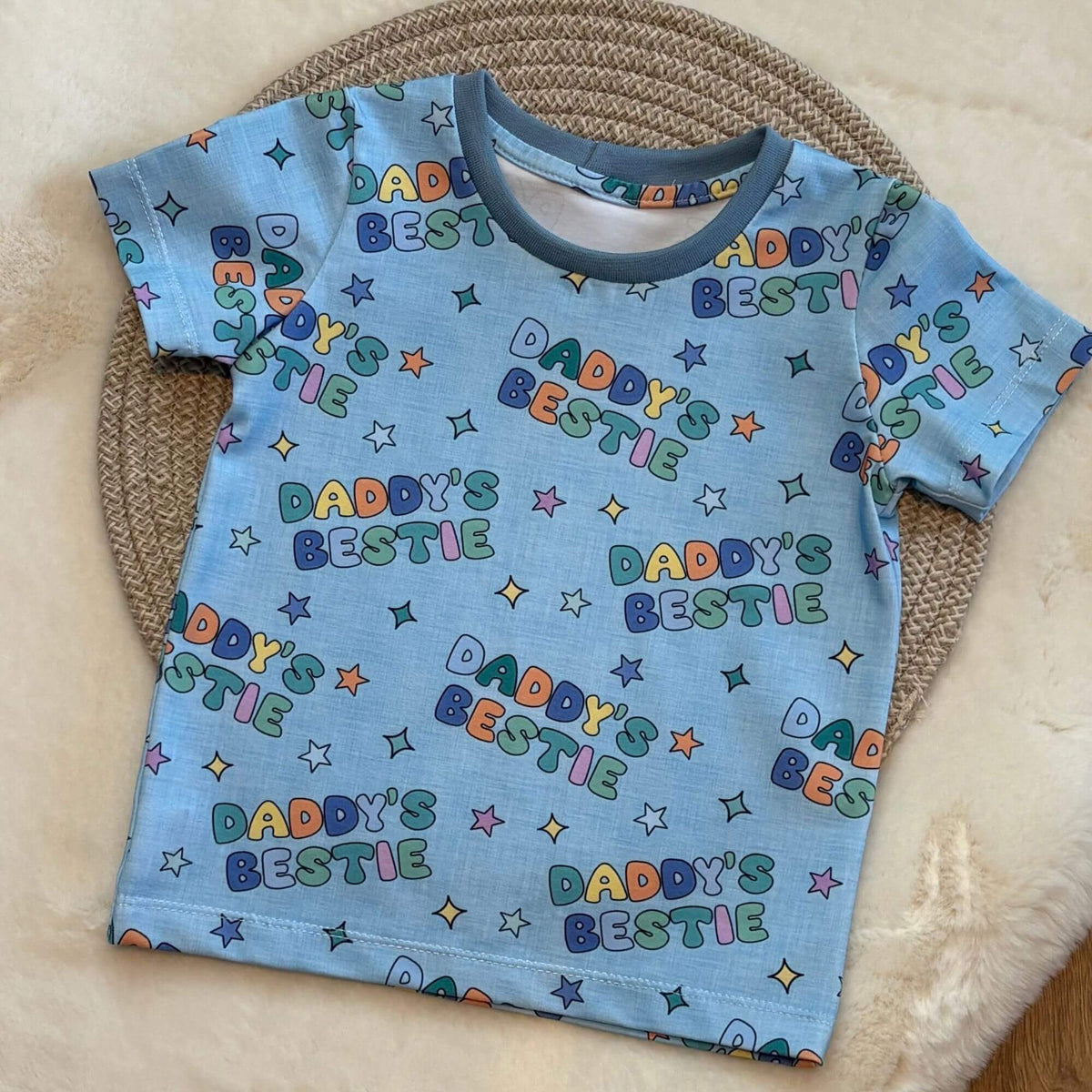 A blue short-sleeve baby t-shirt with an all-over 'Daddy's Bestie' text and star pattern, laid flat on a woven circular mat over a cream faux fur surface.