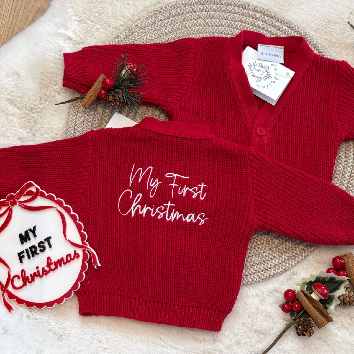 A red ribbed knit baby cardigan sweater shown from both front and back. The back features white cursive embroidery reading 'My First Christmas', and the front is plain red.
