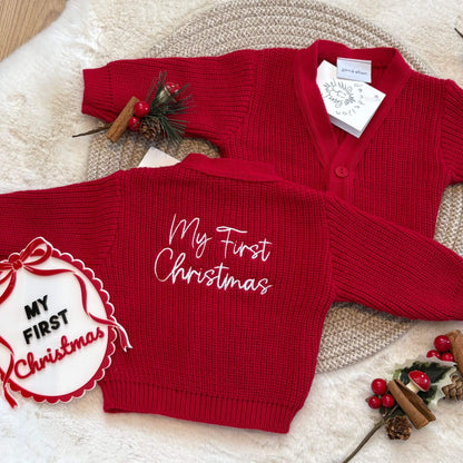 A red ribbed knit baby cardigan sweater shown from both front and back. The back features white cursive embroidery reading 'My First Christmas', and the front is plain red.