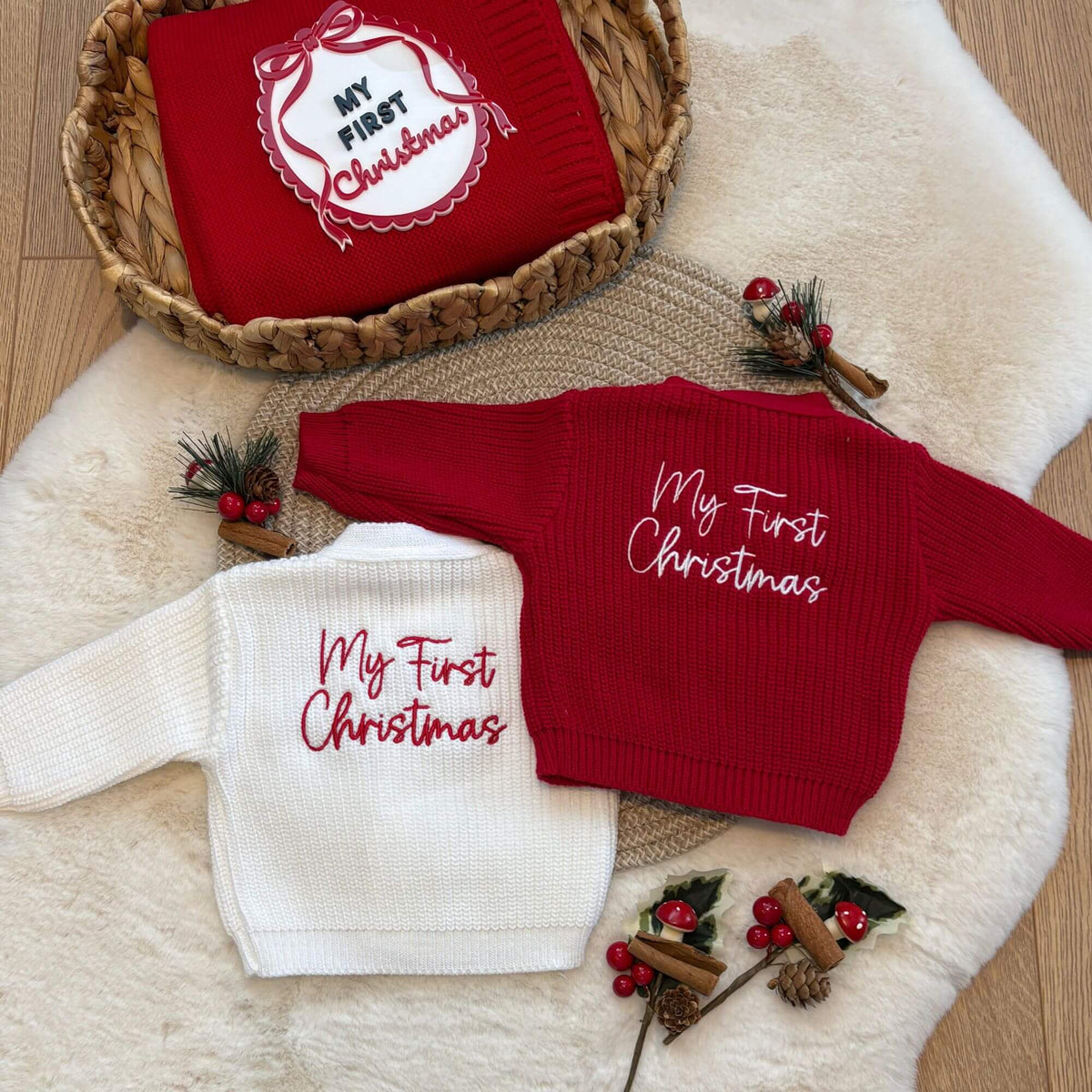 Two personalised baby knit cardigans with 'My First Christmas' embroidery, one in red and one in white, styled on a white faux fur rug with a woven basket containing a matching red knit blanket and acrylic my first Christmas photo prop.