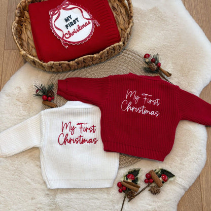 Two personalised baby knit cardigans with 'My First Christmas' embroidery, one in red and one in white, styled on a white faux fur rug with a woven basket containing a matching red knit blanket and acrylic my first Christmas photo prop.