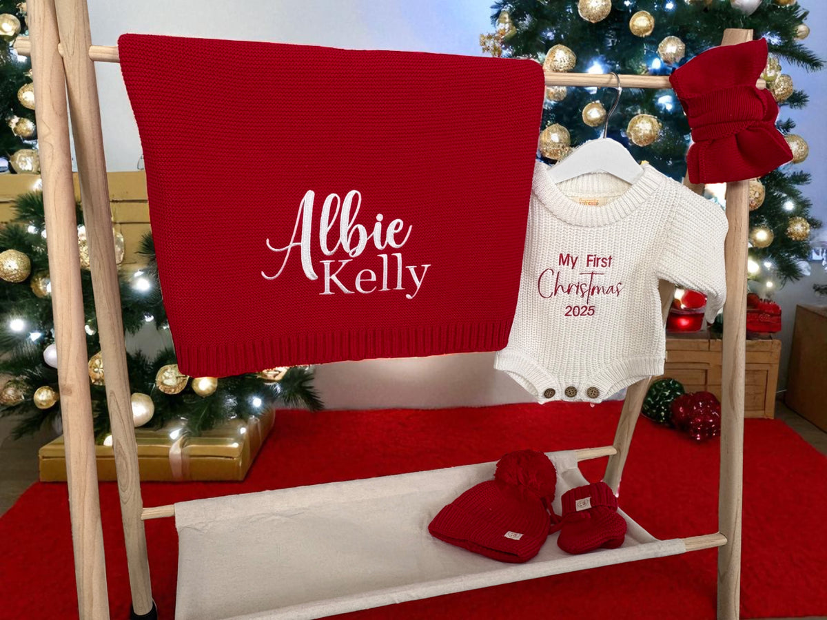 Christmas-themed personalised baby gift set with red blanket, white knit romper, and red booties on a hanger against a decorated Christmas tree background.