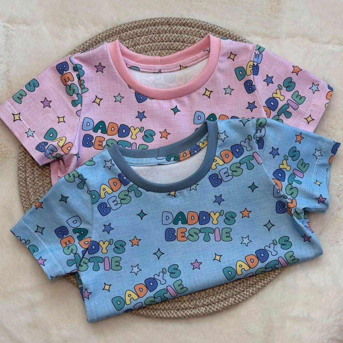 Two short-sleeve baby T-shirts with colorful 'Daddy's Bestie' text and star prints, one in pink and one in light blue, arranged on a woven placemat over a cream faux-fur surface.