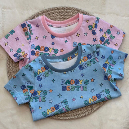 Two short-sleeve baby T-shirts with colorful 'Daddy's Bestie' text and star prints, one in pink and one in light blue, arranged on a woven placemat over a cream faux-fur surface.