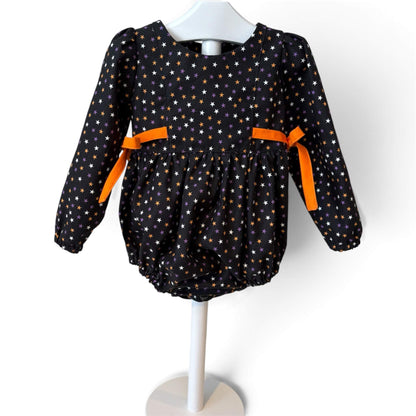 A baby long-sleeve romper in black fabric with a white, orange and purple star pattern, featuring bright orange ribbon bows at the waist.