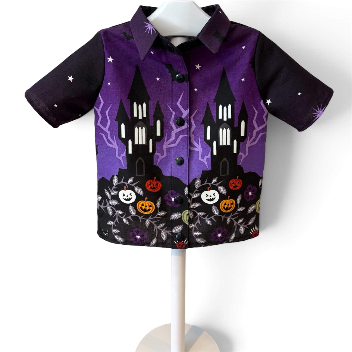 A short-sleeve button-up shirt featuring a Halloween-themed print with purple and black background, castle silhouettes, lightning, pumpkins, flowers, and stars.
