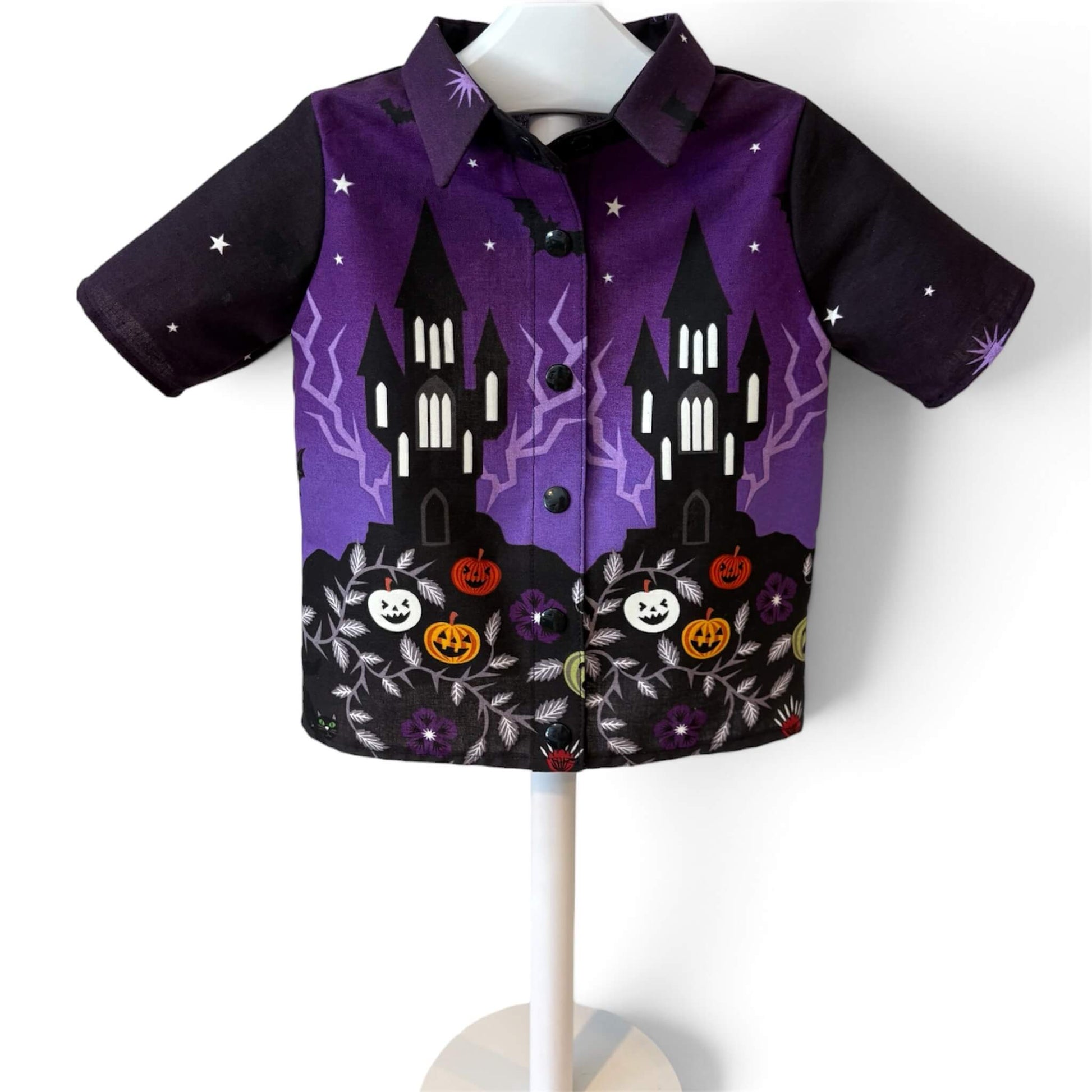 A short-sleeve button-up shirt featuring a Halloween-themed print with purple and black background, castle silhouettes, lightning, pumpkins, flowers, and stars.