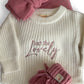Baby sweater with 'Isn't She Lovely' text, pink hat, and booties on a white background