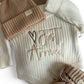 Neutral Knit Newborn Romper Set with “Just Arrived” Embroidery – Includes a cream knit romper, beige pom-pom hat, booties, and matching knit blanket.