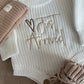 Personally Stitched Baby Knit Outfit – Cream newborn romper with “Just Arrived” embroidered text, paired with beige knit accessories: hat, blanket, and booties.