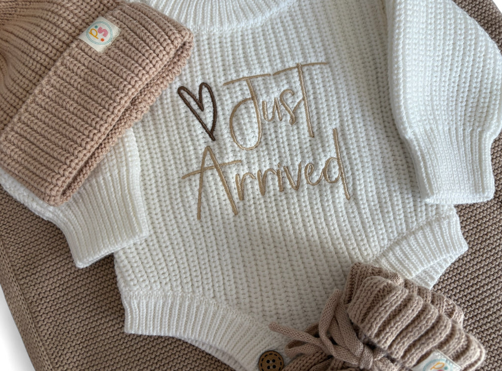 Personally Stitched Baby Knit Outfit – Cream newborn romper with “Just Arrived” embroidered text, paired with beige knit accessories: hat, blanket, and booties.