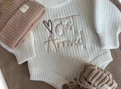 Personally Stitched Baby Knit Outfit – Cream newborn romper with “Just Arrived” embroidered text, paired with beige knit accessories: hat, blanket, and booties.