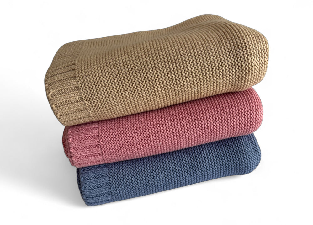 Three stacked knit blankets in beige, pink, and blue on a white background