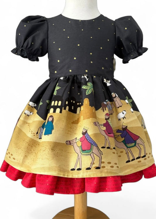 Traditional handmade Christmas dress with nativity print fabric, gold star bow, and red underskirt, available in toddler and girls’ sizes