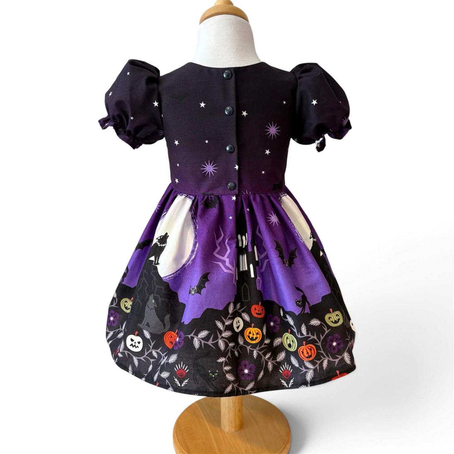 A toddler girl's Halloween dress back view. The dress features a skirt with a moon and wolf silhouette print, and a black hemline decorated with orange jack-o'-lanterns, white skulls, and purple flowers.