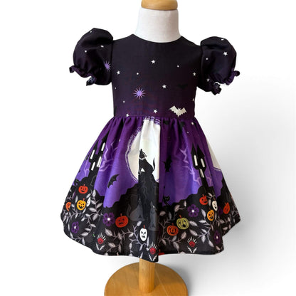 A toddler girl's Halloween dress front view. The dress features a skirt with a moon and wolf silhouette print, and a black hemline decorated with orange jack-o'-lanterns, white skulls, and purple flowers.