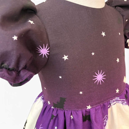A close-up of a children's dress on a mannequin, featuring a dark purple/black bodice with white and lavender starburst prints, puffed sleeves, and a purple skirt with black bat silhouettes and glow in the dark elements