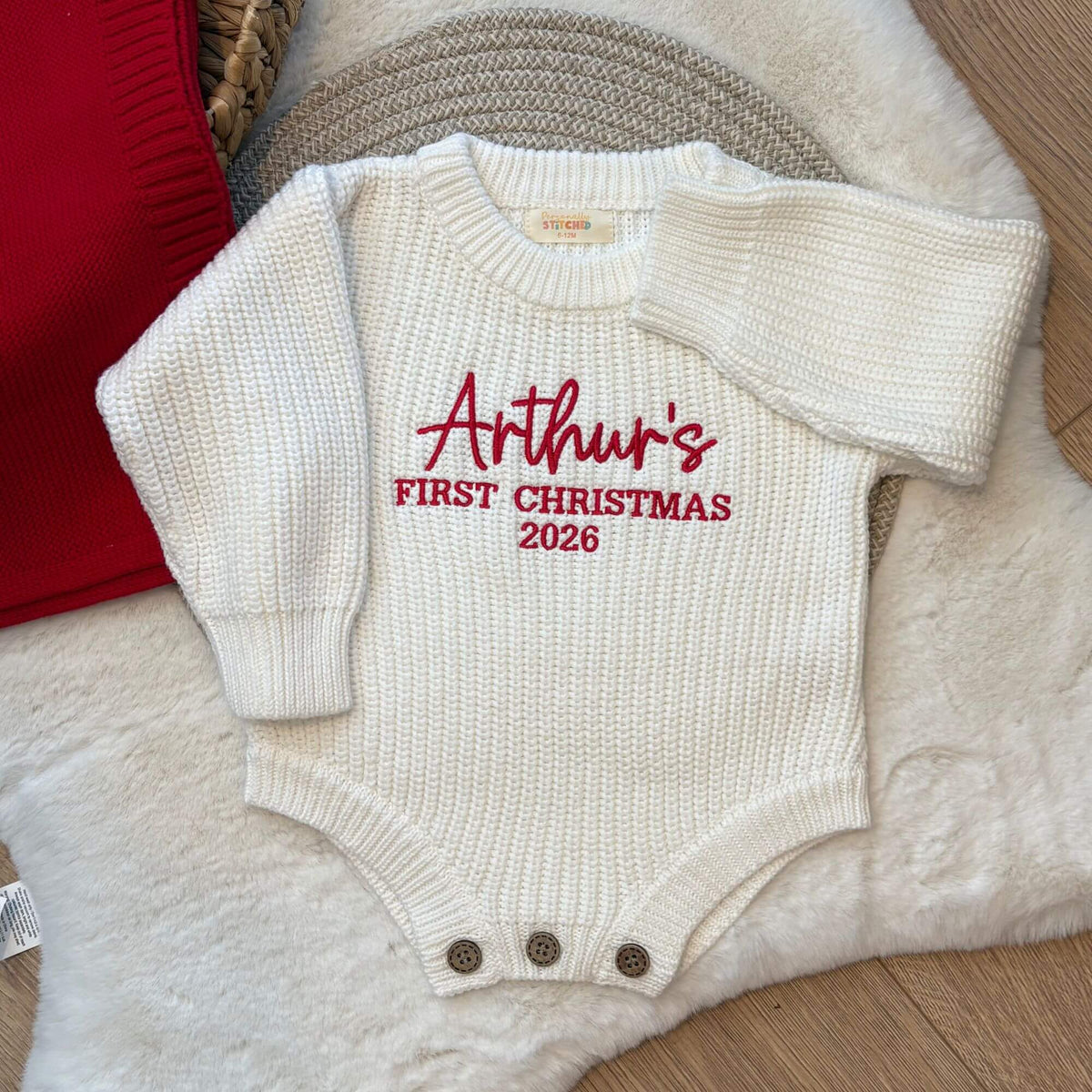 A off white, long-sleeve baby knit bodysuit with 'Arthur's FIRST CHRISTMAS 2026' embroidered in red on the chest, styled on a white faux fur rug with a red blanket that can also be personlaised.