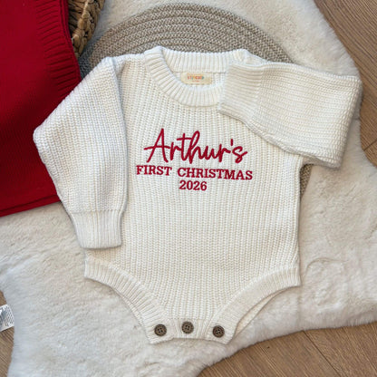 A off white, long-sleeve baby knit bodysuit with 'Arthur's FIRST CHRISTMAS 2026' embroidered in red on the chest, styled on a white faux fur rug with a red blanket that can also be personlaised.