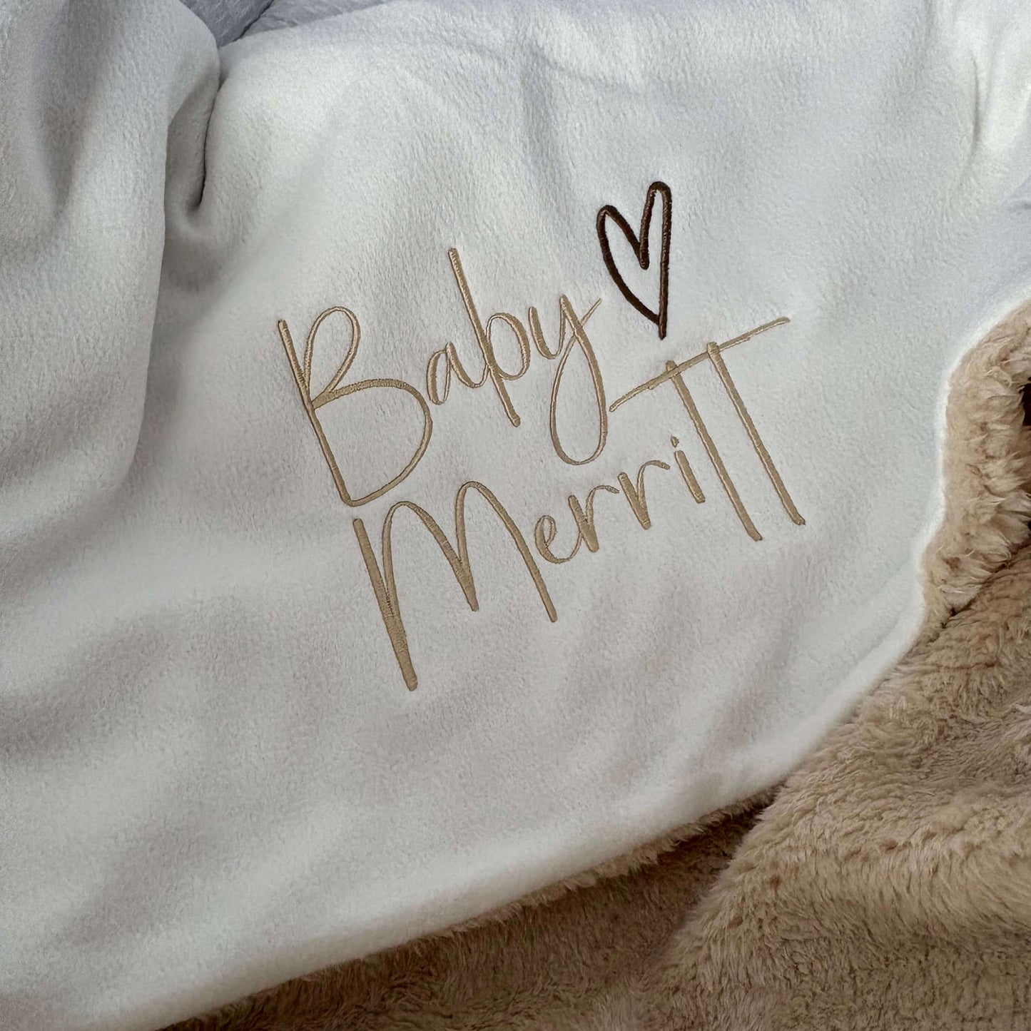 A close-up of a white baby blanket or swaddle with personalised embroidery in a cursive script with a heart symbol, layered over a soft beige fleece blanket.