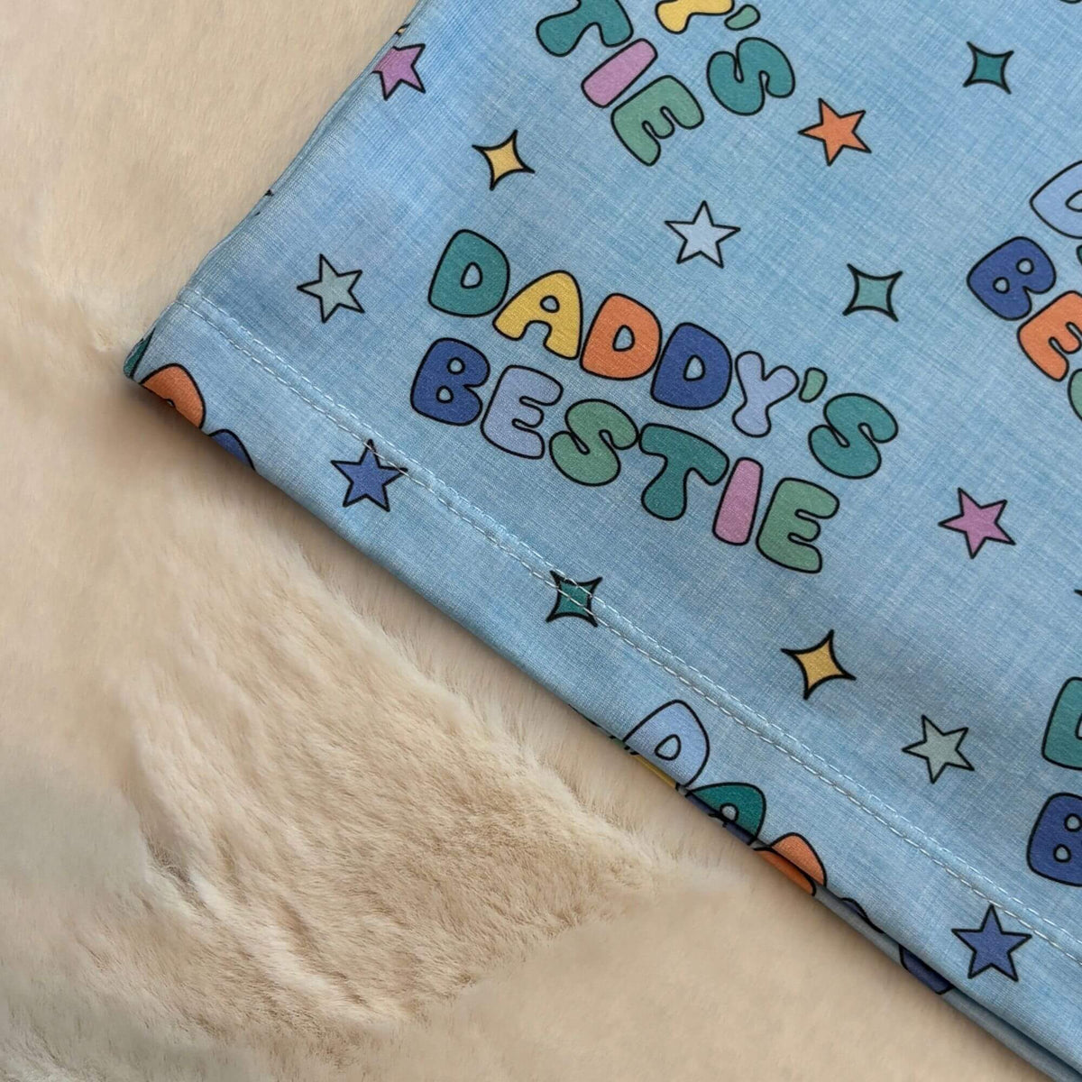 A close-up of a light blue t-shirt printed with colorful retro-style letters spelling 'DADDY'S BESTIE' and small stars, laid on a soft cream faux-fur surface.