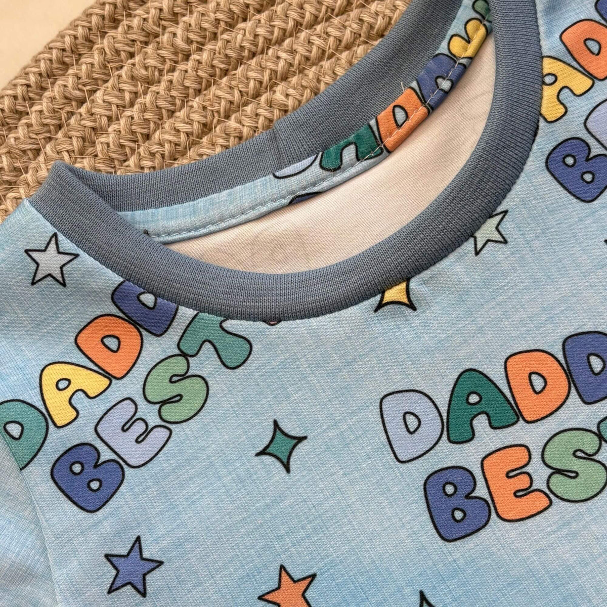 Close-up of a light blue t-shirt with a ribbed crew neck, featuring a colourful 'DADDY'S BEST' text and star pattern, styled on a woven straw mat.
