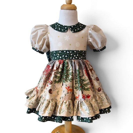 A toddler girl's Christmas-themed dress displayed on a cream dress form. The dress features a cream-colored bodice with white snowflakes, puffy sleeves, a dark green patterned collar and waistband, and a tiered skirt with a festive print of Christmas trees, Hedgehogs, snowmen and red ornaments.