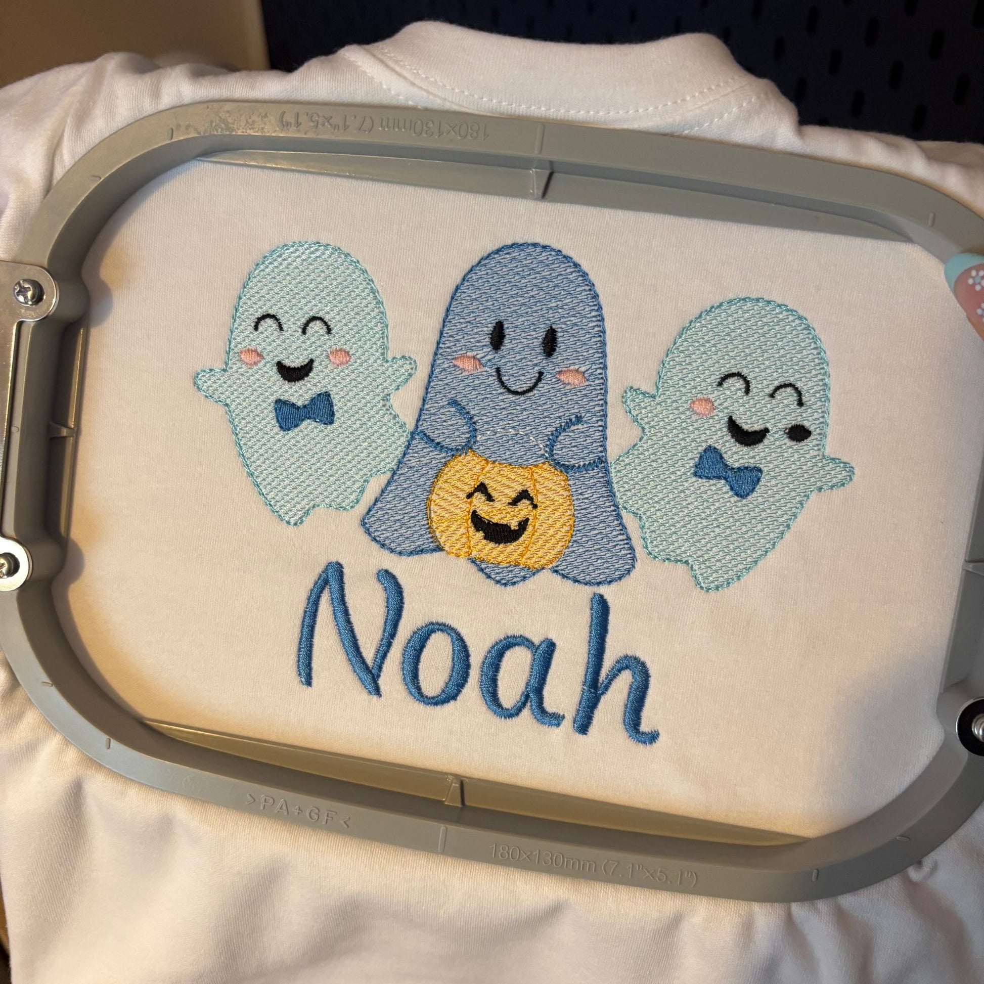 A close-up of a custom embroidery design held in a hoop, featuring three smiling ghosts in light blue and medium blue with a yellow pumpkin in the center, and the name 'Noah' embroidered below in blue.