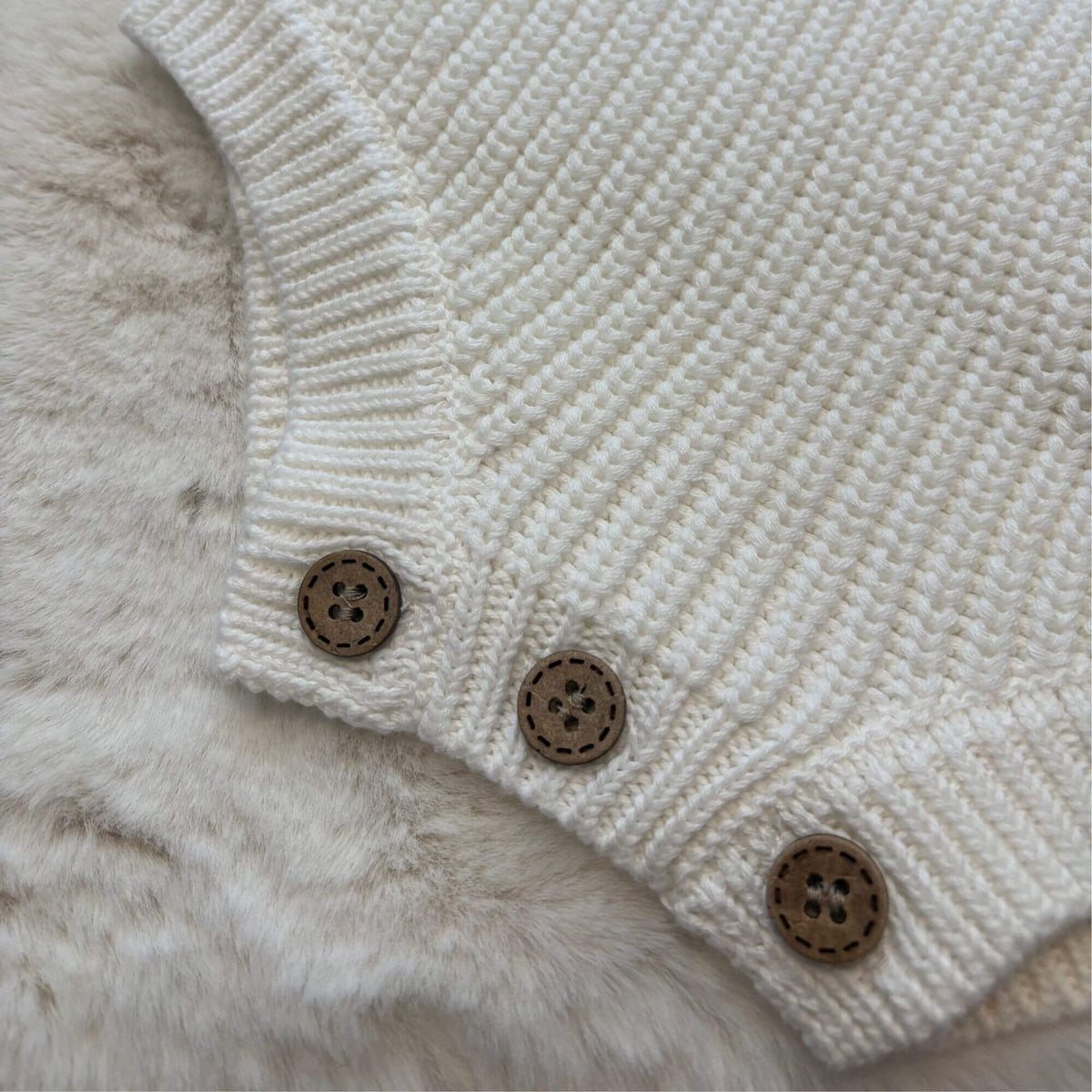 Close-up of a white-coloured knit baby romper with three brown wooden buttons, laid flat on a soft white faux fur surface.