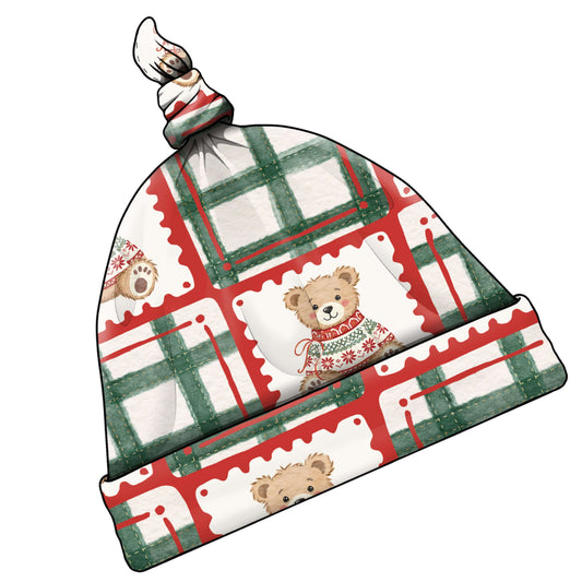 A knotted baby hat with a festive holiday print featuring red and green plaid stripes, white stamp-like squares, and cartoon teddy bears wearing Christmas sweaters.