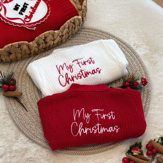 Two folded knit cardigans displayed on a woven placemat: a red sweater with white embroidered text reading 'My First Christmas' and a white sweater with red embroidered text reading 'My First Christmas.' A red woven blanket with a holiday design  acrylic photo prob is partially visible at the top left, and small holiday props including berries, pinecones, cinnamon sticks, and greenery surround the sweaters.