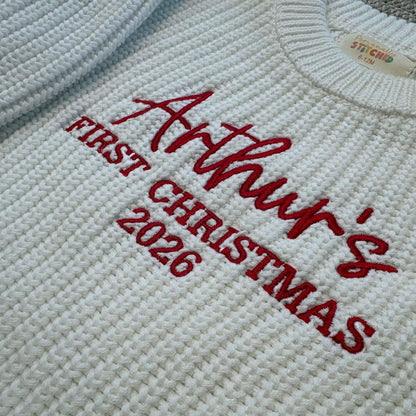 Close-up of a white knit sweater with red embroidered text reading 'Arthur's First Christmas 2026' on the chest.