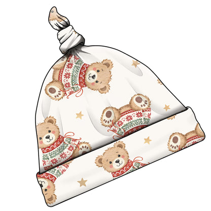 A knotted baby beanie hat featuring a repeating pattern of cute teddy bears wearing festive red and green knit sweaters, with small gold stars on an off-white background.