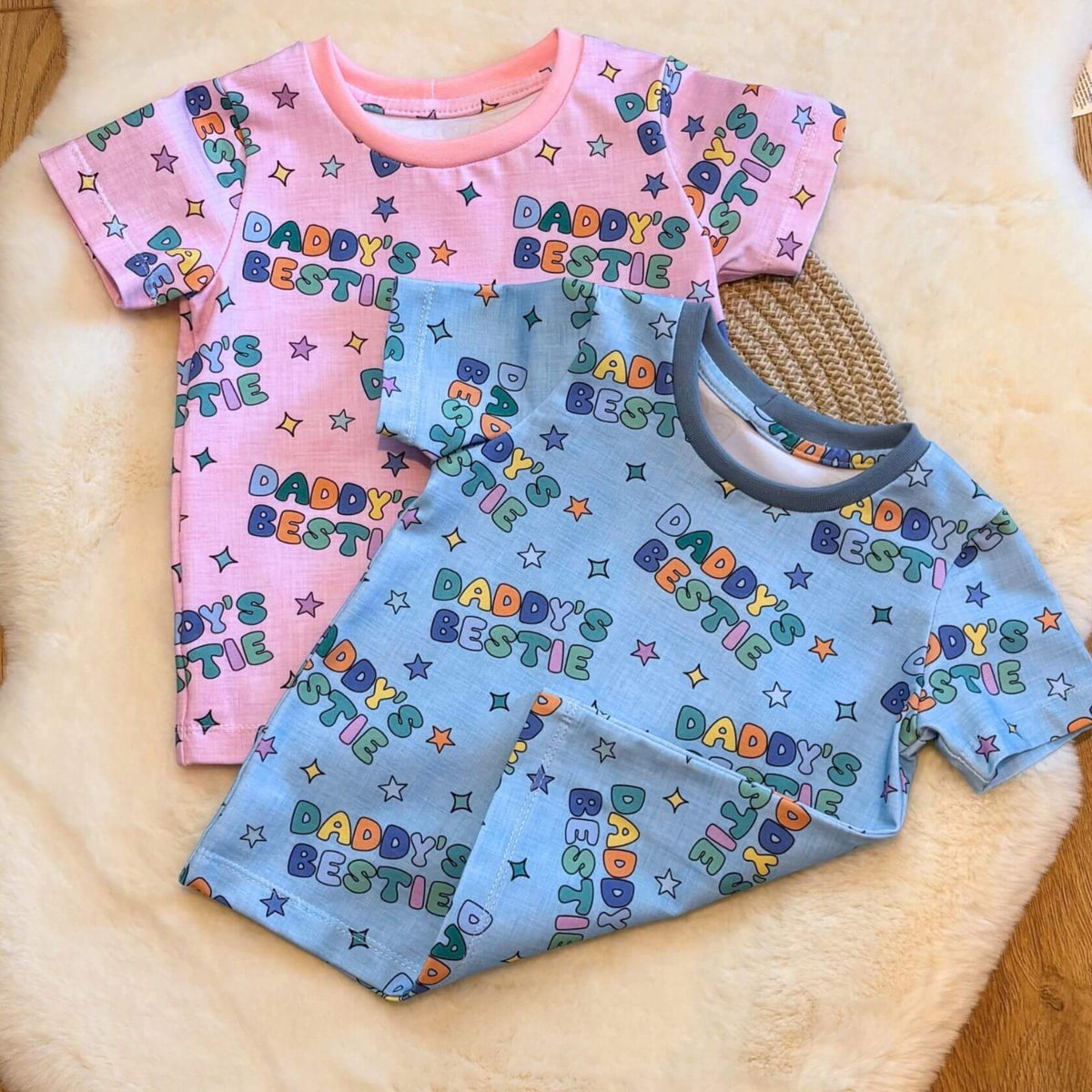 Two short-sleeve baby tshirts laid flat on a cream faux-fur rug over a wood floor. One set is light pink with multicolor 'DADDYY'S BESTIE' text and star motifs, and the other is light blue with the same print.