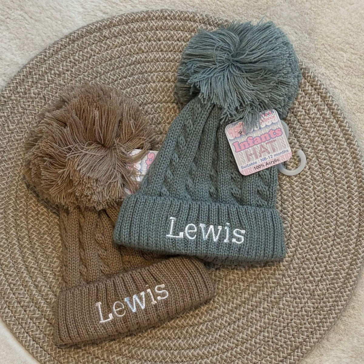 Two cable-knit baby beanies with large pom-poms and embroidered text, displayed on a woven round mat. One beanie is coffee brown and the other is sage green.