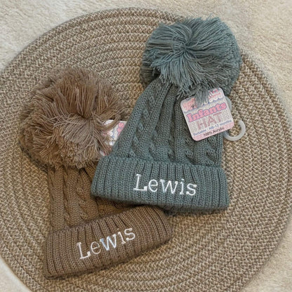 Two cable-knit baby beanies with large pom-poms and embroidered text, displayed on a woven round mat. One beanie is coffee brown and the other is sage green.