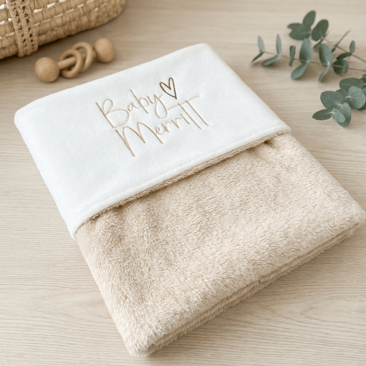 A folded baby blanket or wrap in soft cream and beige tones, with the top section in white embroidered with babies name in a warm matching coloured thread. The blanket is styled on a light wood surface with a woven basket, wooden teething rings, and eucalyptus leaves in the background.