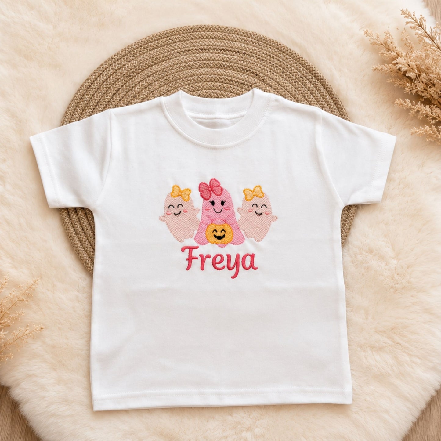 A white short-sleeve kids t-shirt laid flat. The shirt features a graphic of three cute pink ghosts with orange bows and a smiling yellow pumpkin, with the name  embroidered in pink below them.