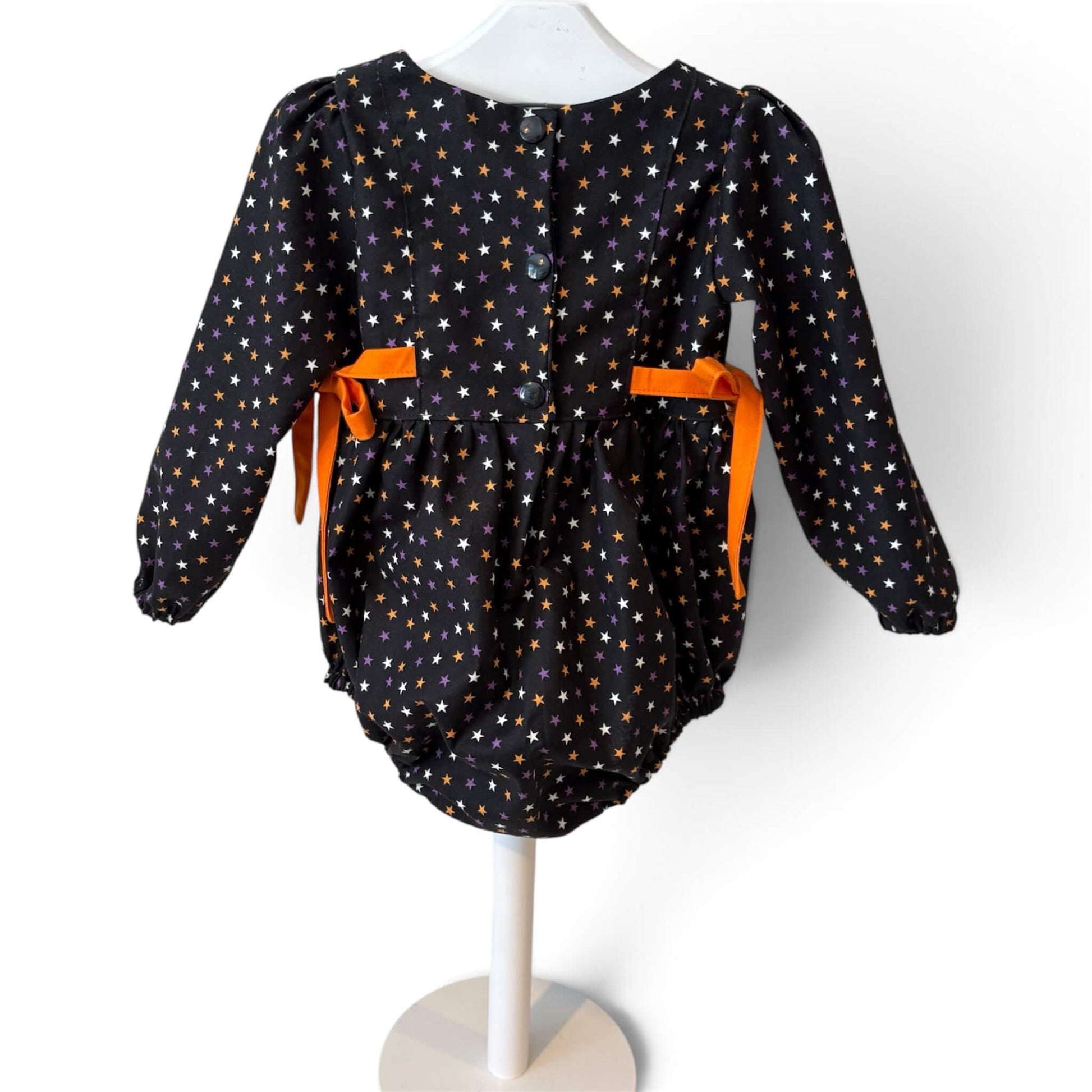 A back view of a baby romper in black with an all-over pattern of small white, orange and purple stars, featuring bright orange ribbon ties at the waist.