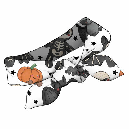 A white fabric hair bow with a Halloween-themed print featuring gray bats, a smiling orange pumpkin, black spiderwebs, and black stars.