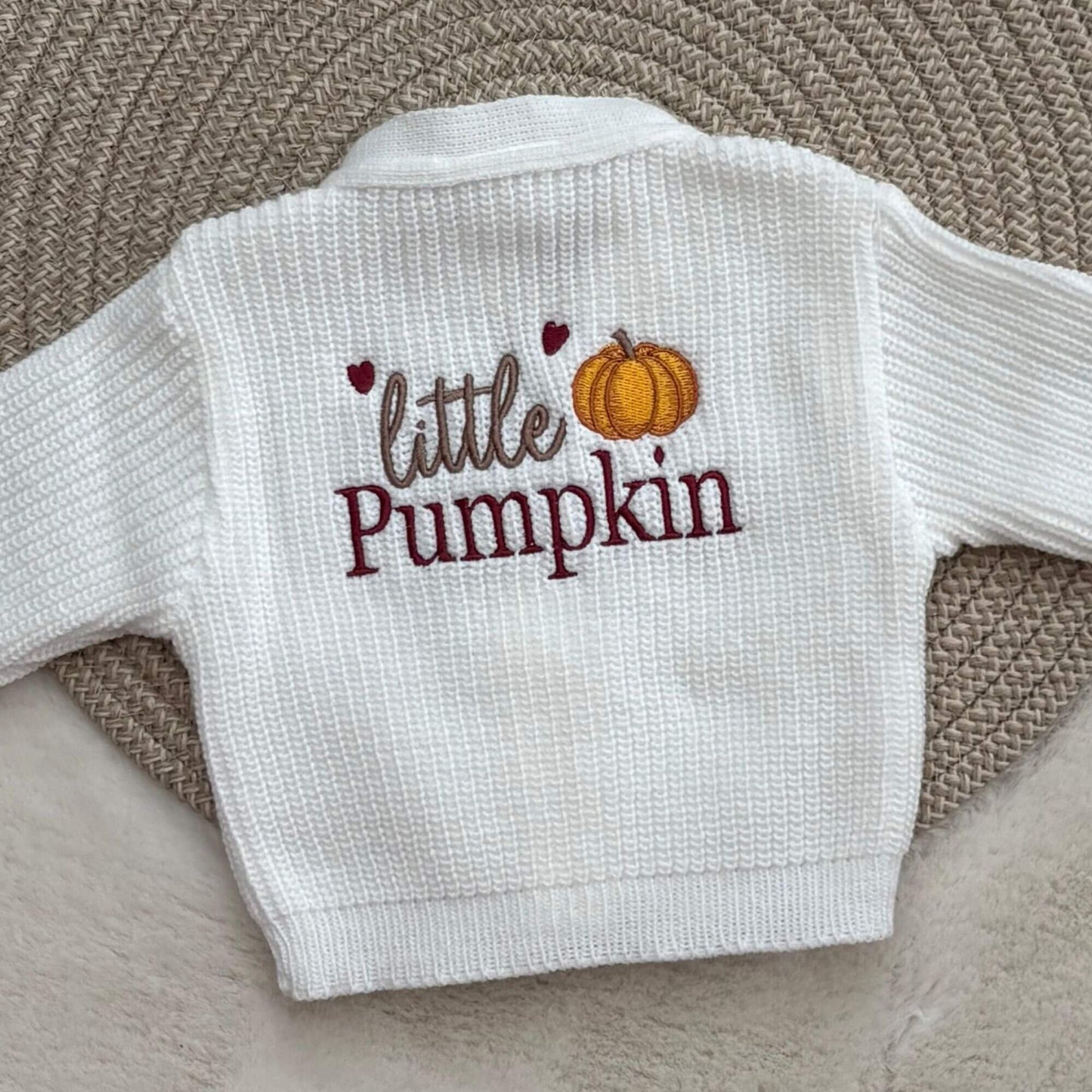 A white ribbed knit baby cardigan featuring the words 'little Pumpkin' in brown and burgundy embroidery with small  hearts and an orange pumpkin graphic on the back.